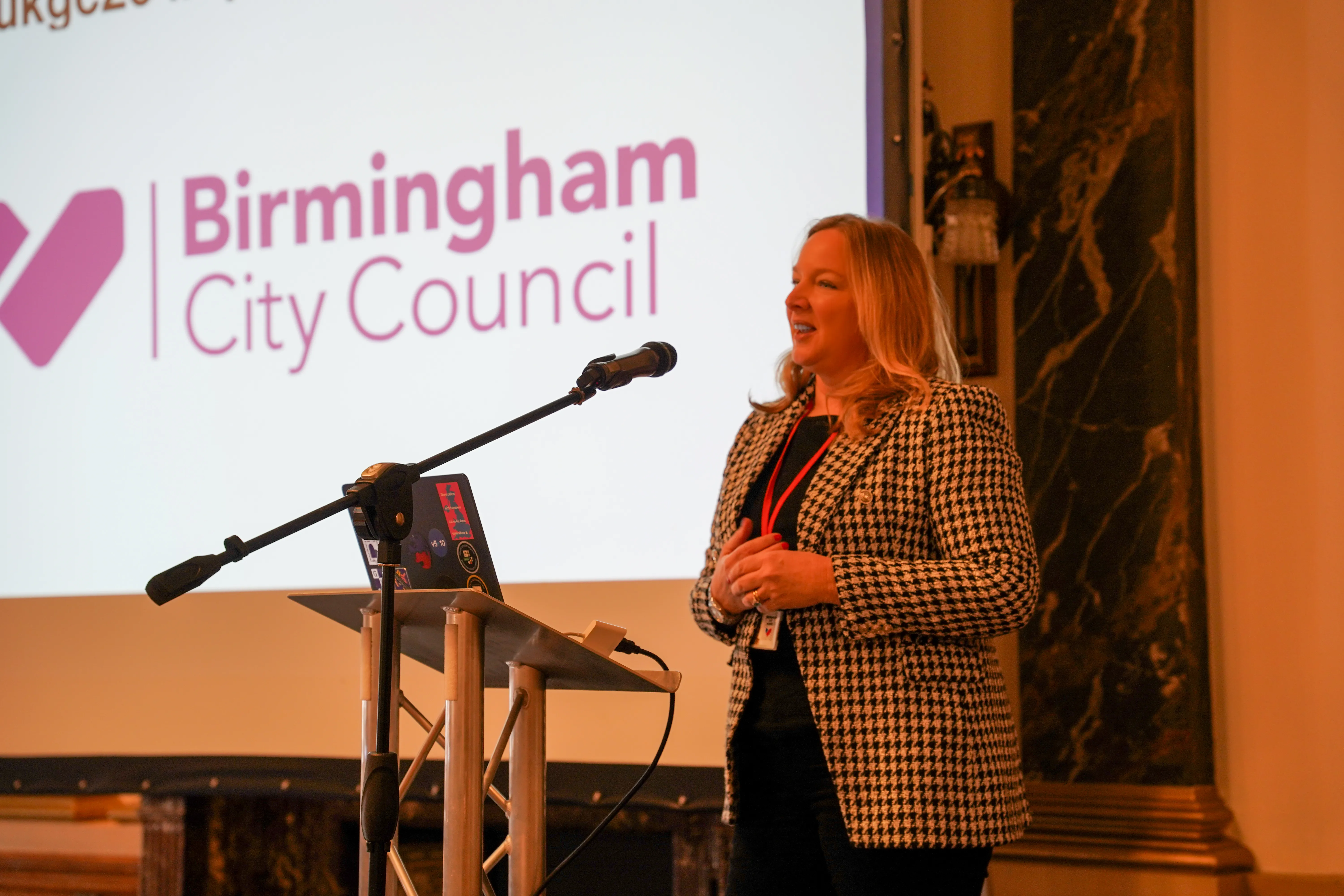 Katy Fox FCIPD, CPO of Birmingham City Council, wearing a grey checked suit, stands on a stage with a microphone, smiling and gesturing to welcome a large audience in a brightly lit hall.