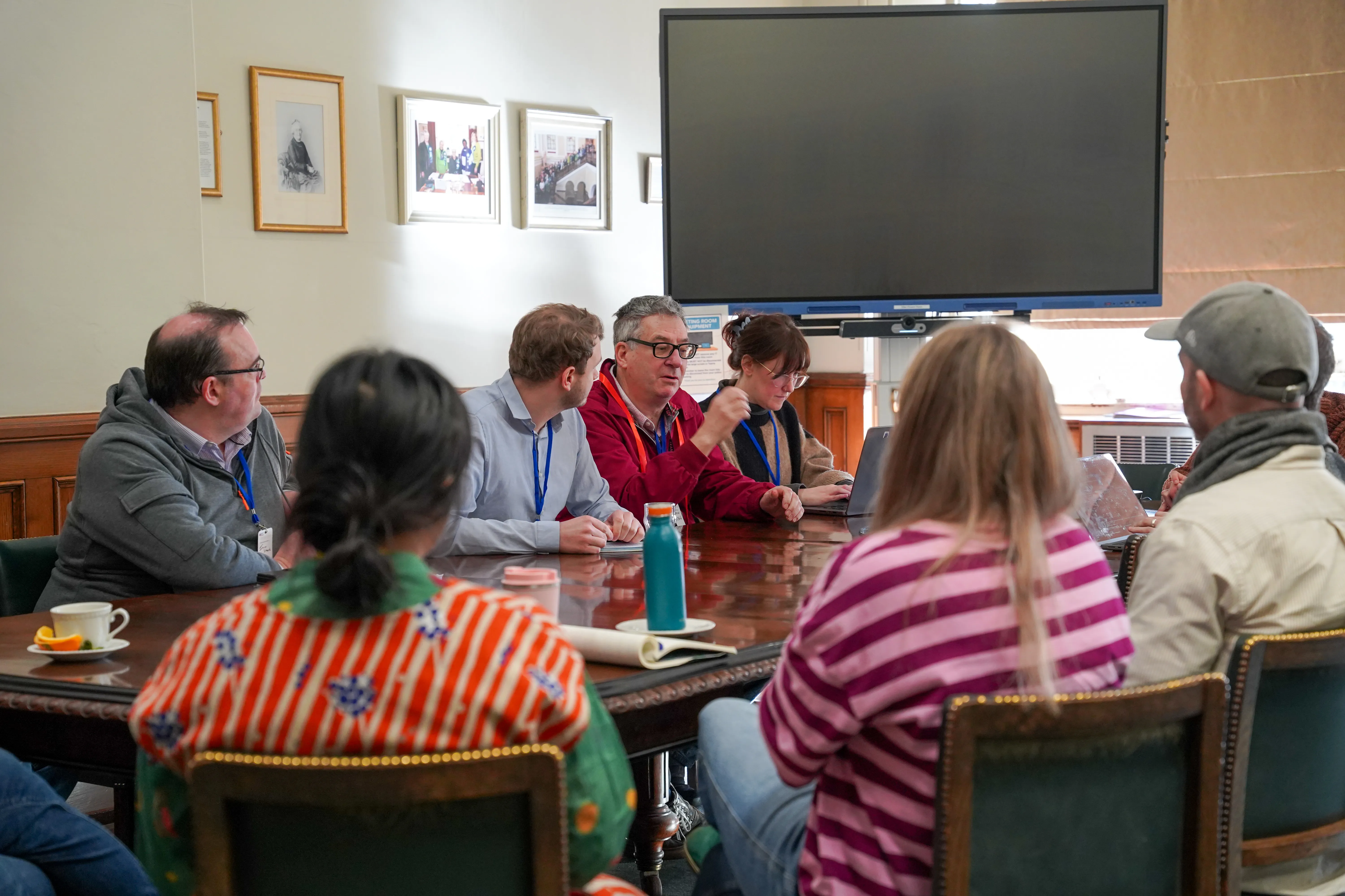 A group of seven diverse participants sitting around a circular wooden table in a brightly lit office, engaged in an active discussion with notebooks and laptops open.