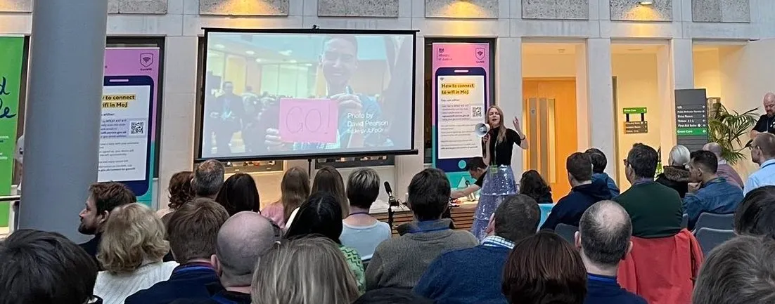 A picture of the opening address at UKGovcamp 2023, the seated audience watching Amanda Smith introducing the event through a megaphone.